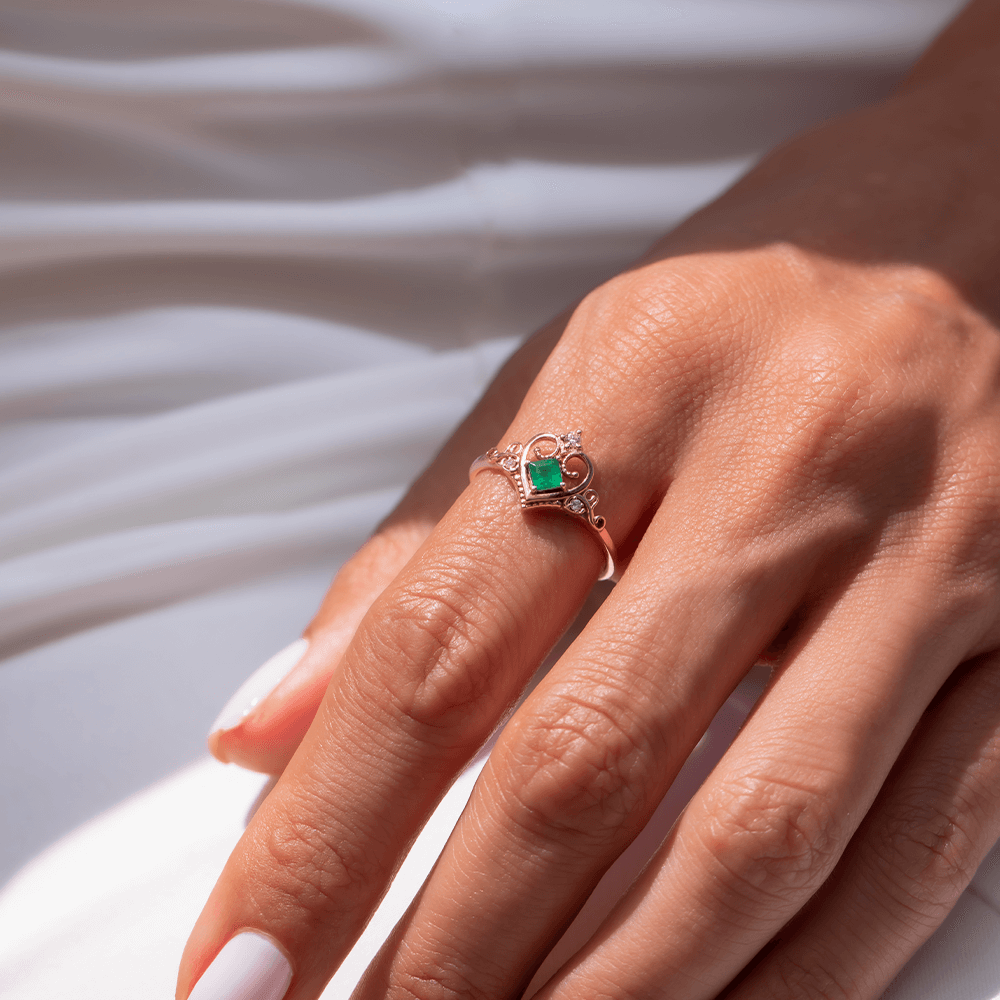 ortasında kare kesim zümrüt taşı ve zümrüt taşının üzerinde ve yanlarında bulunan topam 3 adet küçük pırlanta taşlarıyla tasarlanmış vintage tarzı altın taç yüzüğün kadın modelin parmağındaki görüntüsü