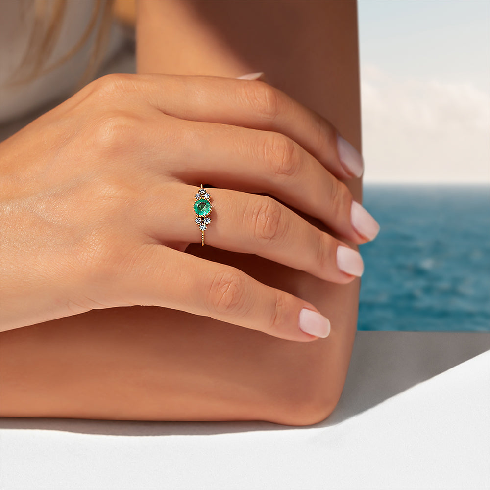 kenarlarında pırlantalar bulunan ve ortasında yuvarlak kesim zümrüt taşı bulunan yüzüğü takmış bir kadın modelin kolunu tutarak yüzüğünü gösterdiği bir pozu